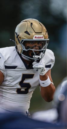 A person wearing a gold football helmet and dark uniform is focused on the game while another person in a white uniform is partially visible. Description generated by AI