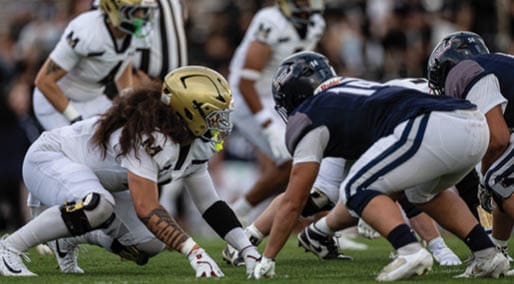 Two people in football uniforms are engaged in a play on the field, with one person reaching out toward the other near the ground. Description generated by AI