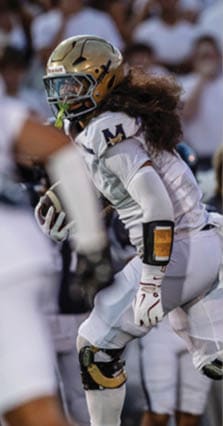 A person wearing a dark sports uniform with the number six on the back is running on a field while another person in white sports gear extends a leg toward them. Description generated by AI