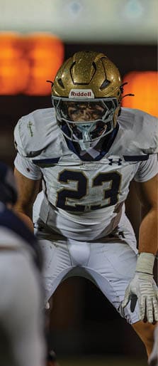 A person wearing a gold football helmet and white uniform with the number 23 is in a ready stance on a football field at night. Description generated by AI