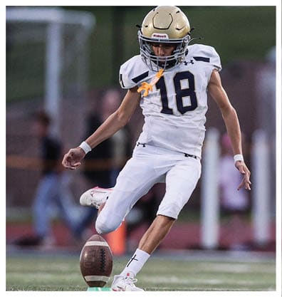 A person wearing a white football uniform is kicking a football on a field during a game. Description generated by AI