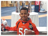 A child wearing an orange Broncos football jersey is smiling while holding a football and a stuffed horse toy in a room with trophies and sports equipment in the background. Description generated by AI