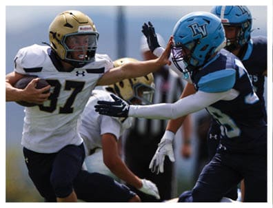 Two people in football uniforms are engaged in a play with one holding the ball and the other reaching out to block or tackle. Description generated by AI