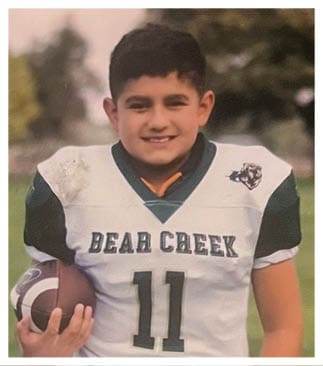 A child wearing a Bear Creek football jersey is smiling outdoors with trees and grass in the background. Description generated by AI