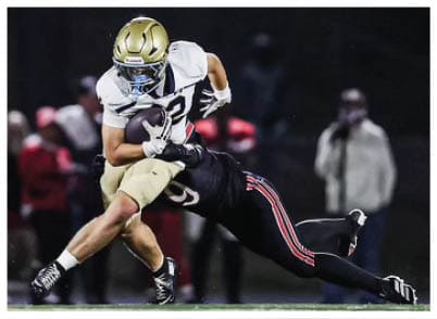 A person wearing a gold helmet and white football uniform is running with a football on a dark field. Description generated by AI