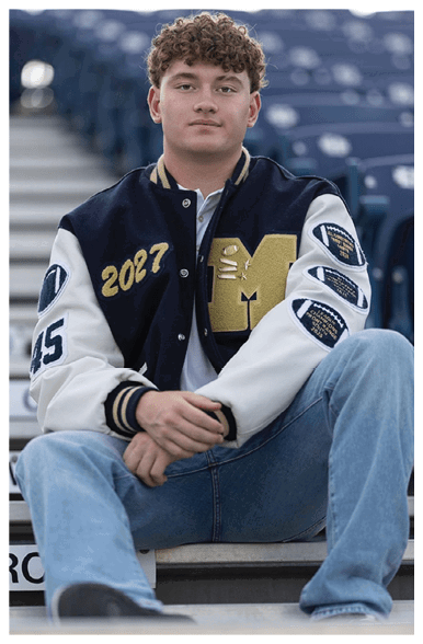 A person with curly hair wearing a letterman jacket and jeans is sitting on stadium bleachers with hands clasped and looking forward. Description generated by AI
