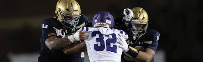 Three people in football uniforms are engaged in a play on a field, with two players in dark uniforms tackling a player in a white jersey numbered 32. Description generated by AI