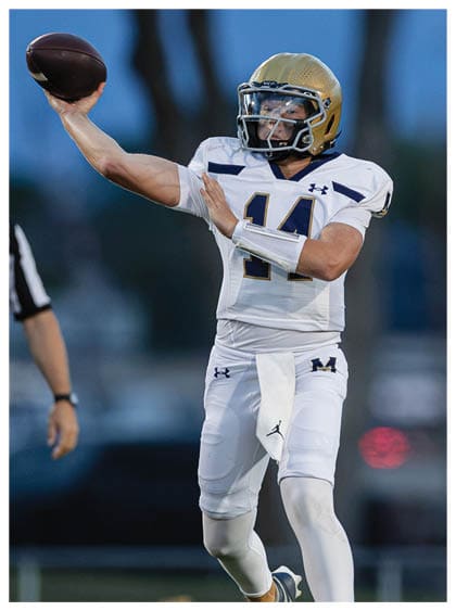 A person wearing a white football uniform with the number 14 and a gold helmet is throwing a football on a field during a game. Description generated by AI