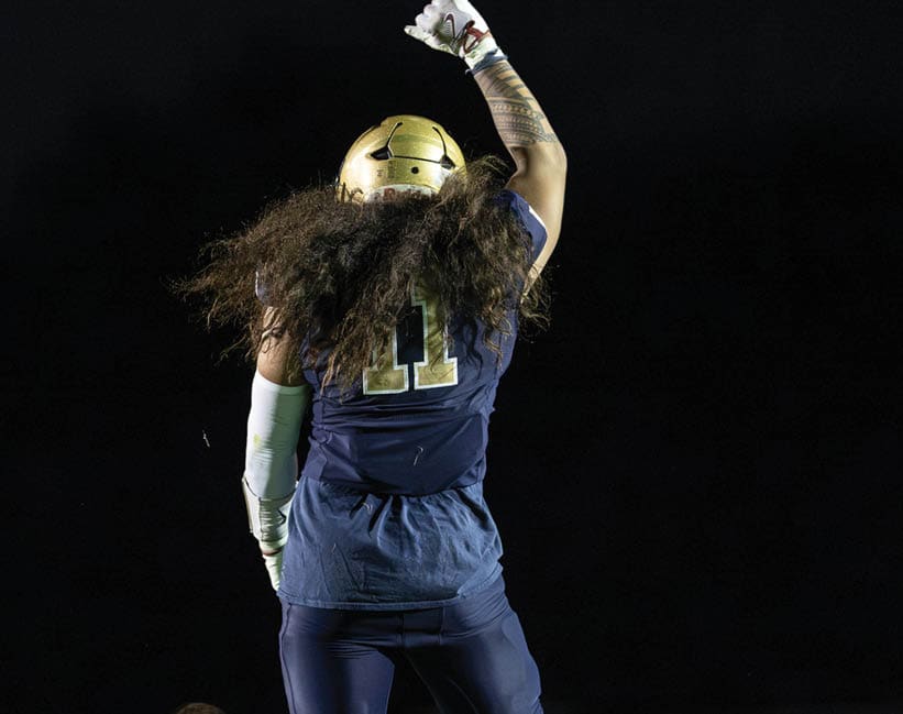 A person wearing a gold helmet and dark blue sports uniform with the number 11 raises one arm while standing on a field at night. Description generated by AI