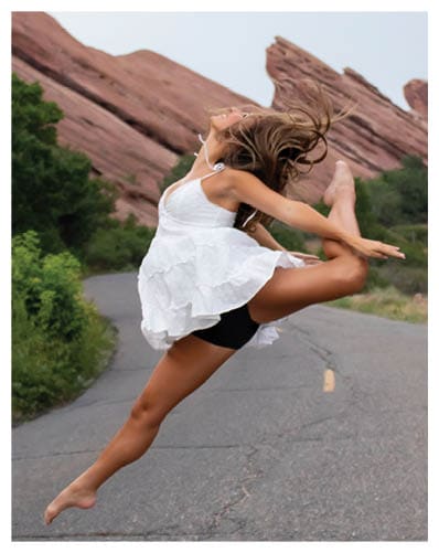 A person in a white dress is captured mid-jump with hair flowing against a backdrop of large red rock formations and greenery. Description generated by AI