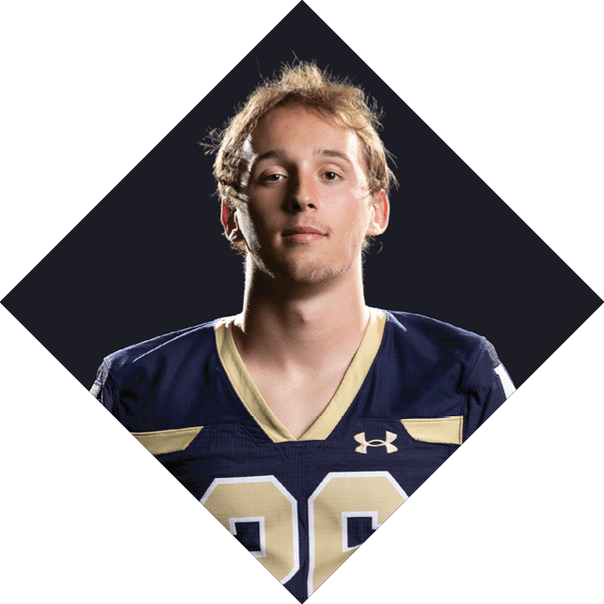 A person with light curly hair wearing a dark blue and gold sports jersey with the number 26 stands against a dark background. Description generated by AI