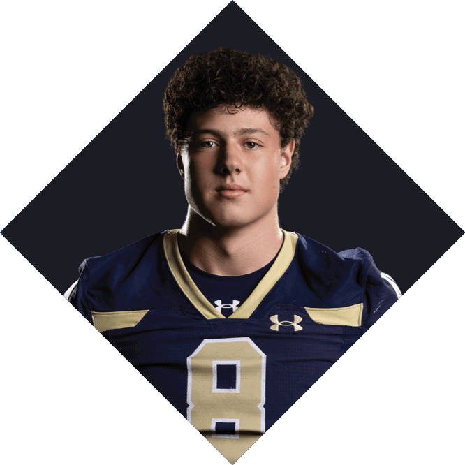 Person with curly hair wearing a dark blue football jersey with the number eight stands against a dark background. Description generated by AI