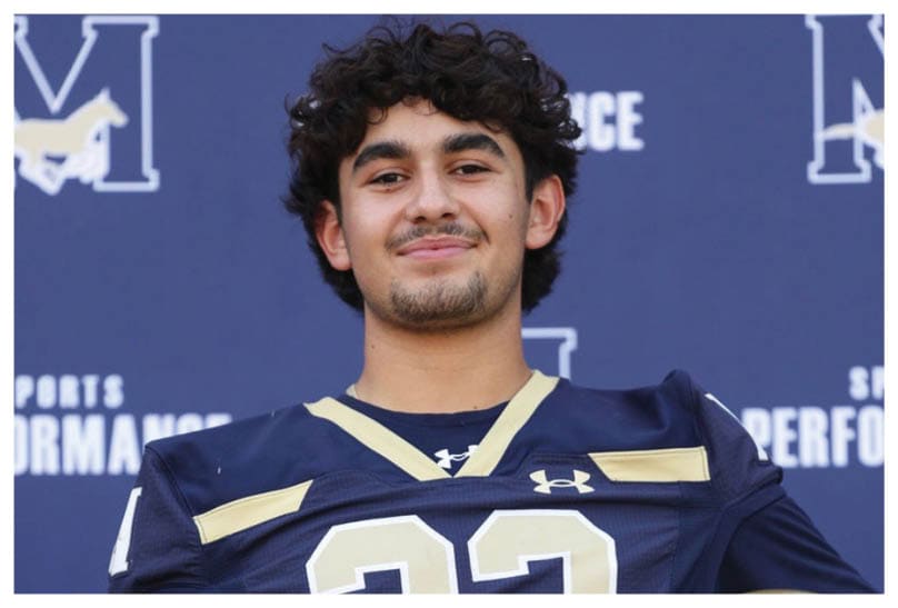 A person wearing a dark blue football jersey with the number 22 is holding a gold football helmet and standing in front of a backdrop with sports performance logos. Description generated by AI