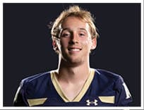 A person with short, light-colored hair is smiling and wearing a dark blue sports jersey with gold accents against a black background. Description generated by AI
