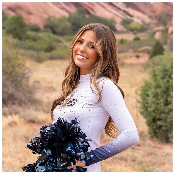 A person with long hair wearing a white top is smiling outdoors with large red rock formations and greenery in the background. Description generated by AI