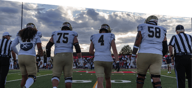 Four people wearing sports gear and cleats are walking on a football field near yard lines. Description generated by AI