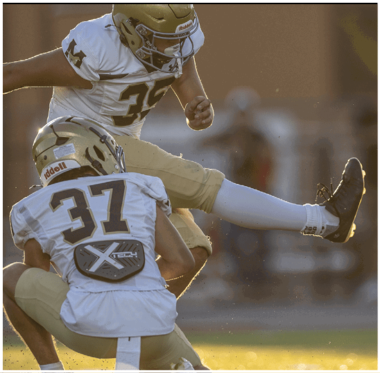 Two people in football uniforms are engaged in a play with one person kicking a ball while the other is positioned in front wearing a helmet and protective gear. Description generated by AI