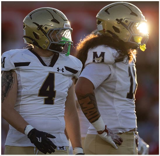 Two people wearing football uniforms with gloves and tattoos are standing side by side on a field. Description generated by AI