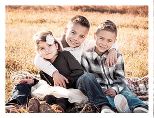 Three children sitting closely together on grass in a sunlit outdoor setting, with one child wearing a headband with a flower and the others dressed in casual sweaters and jeans. Description generated by AI