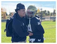 Two people on a football field are smiling and embracing, with one wearing a football uniform and helmet and the other dressed casually with a backpack and cap. Description generated by AI