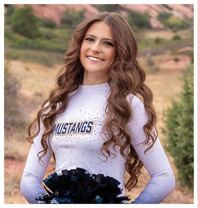 A person with long wavy hair wearing a white Mustangs shirt is smiling outdoors with rocky hills and greenery in the background. Description generated by AI