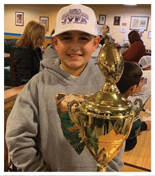 A child wearing a gray hoodie with a bear graphic and a white cap is smiling while holding a large gold trophy shaped like a football. Description generated by AI