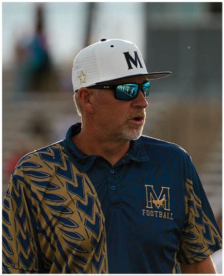 A person wearing a white and black cap with the letter M, reflective sunglasses, and a blue and gold patterned sports shirt is looking to the side outdoors. Description generated by AI