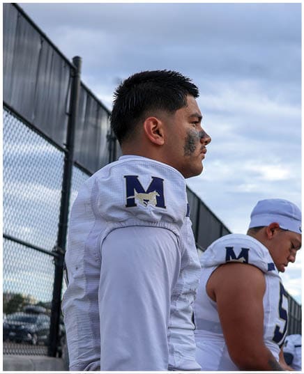 A person wearing a white sports jersey with a large letter M and a mustang logo on the shoulder stands near a chain-link fence under a cloudy sky. Description generated by AI