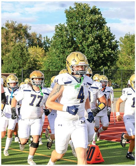A group of people wearing white and gold football uniforms and helmets are standing outdoors on a field with trees and a partly cloudy sky in the background. Description generated by AI