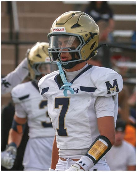 A person wearing a gold football helmet and white jersey with a blue “M" stands on a field with blurred spectators in the background. Description generated by AI