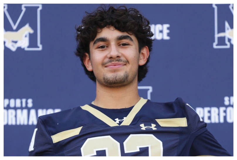 A person wearing a dark blue football jersey with the number 22 is holding a gold football helmet and standing in front of a backdrop with sports performance logos. Description generated by AI