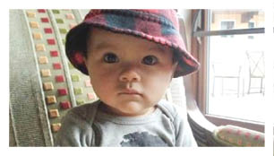 A child wearing a plaid bucket hat and a gray shirt with a printed face design is sitting on a patterned chair near a window. Description generated by AI