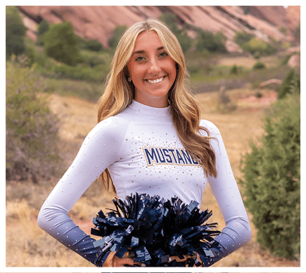 A person with long hair wearing a white and blue cheerleading uniform holding blue and black pom-poms stands outdoors with rocky hills and greenery in the background. Description generated by AI