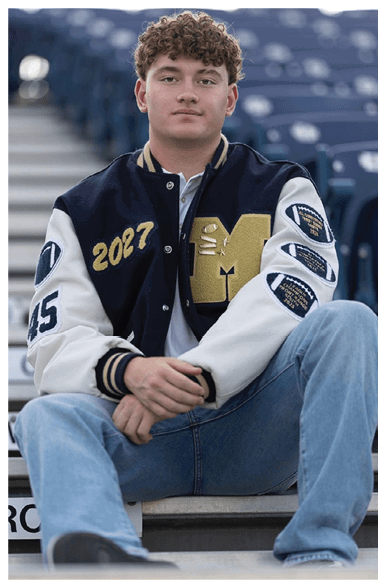 A person with curly hair wearing a letterman jacket and jeans is sitting on stadium bleachers with hands clasped and looking forward. Description generated by AI