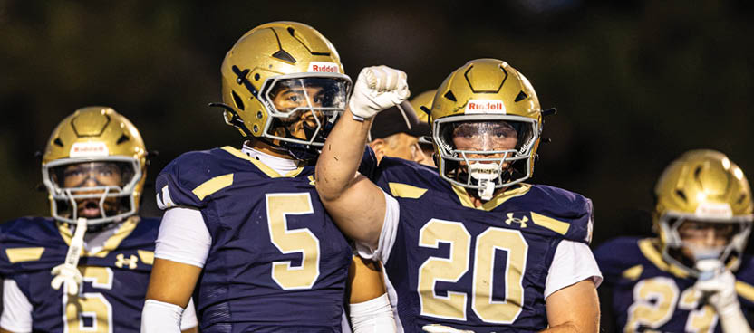 Three football players in gold helmets and navy blue uniforms stand together on a field, with one player raising a fist in celebration. Description generated by AI