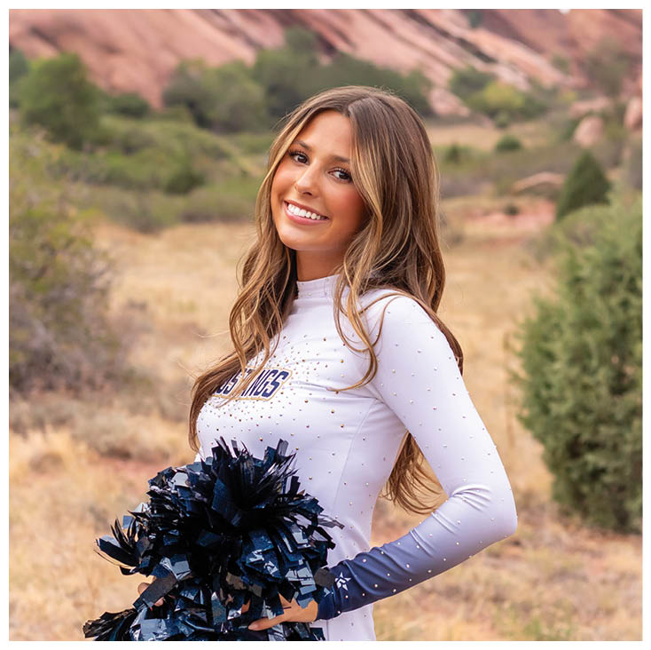 A person with long hair wearing a white top is smiling outdoors with large red rock formations and greenery in the background. Description generated by AI