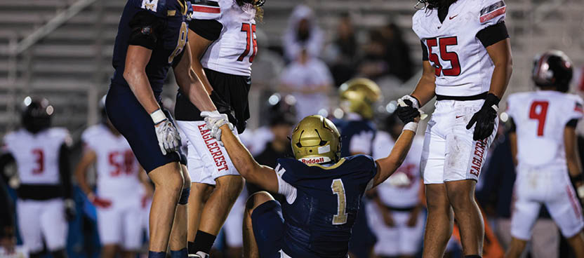 A person wearing a football uniform and helmet is sitting on the ground while two other people in football uniforms help them up on a grassy field during a game. Description generated by AI