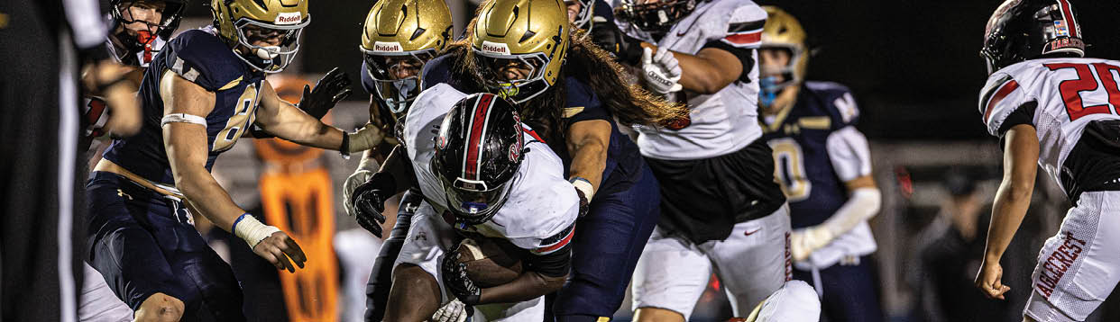 Several people in football uniforms are engaged in a tackle during a nighttime game on a field. Description generated by AI