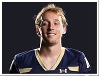 A person with short, light-colored hair is smiling and wearing a dark blue sports jersey with gold accents against a black background. Description generated by AI