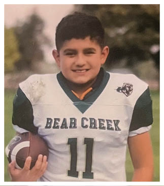 A child wearing a Bear Creek football jersey is smiling outdoors with trees and grass in the background. Description generated by AI