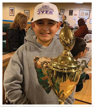 A child wearing a gray hoodie with a bear graphic and a white cap is smiling while holding a large gold trophy shaped like a football. Description generated by AI