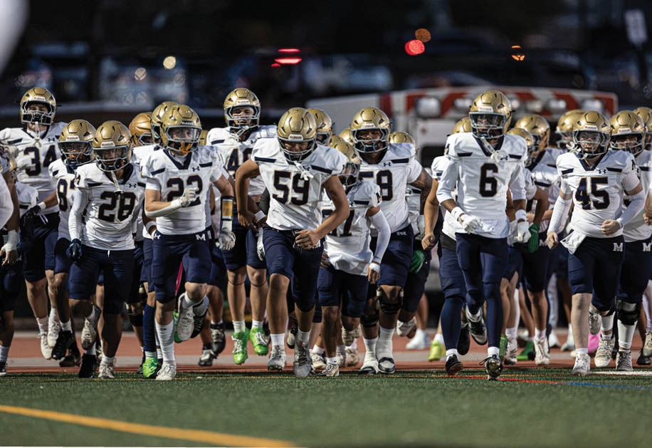 A group of football players in white and navy uniforms with gold helmets are running together on a field during a game or practice. Description generated by AI