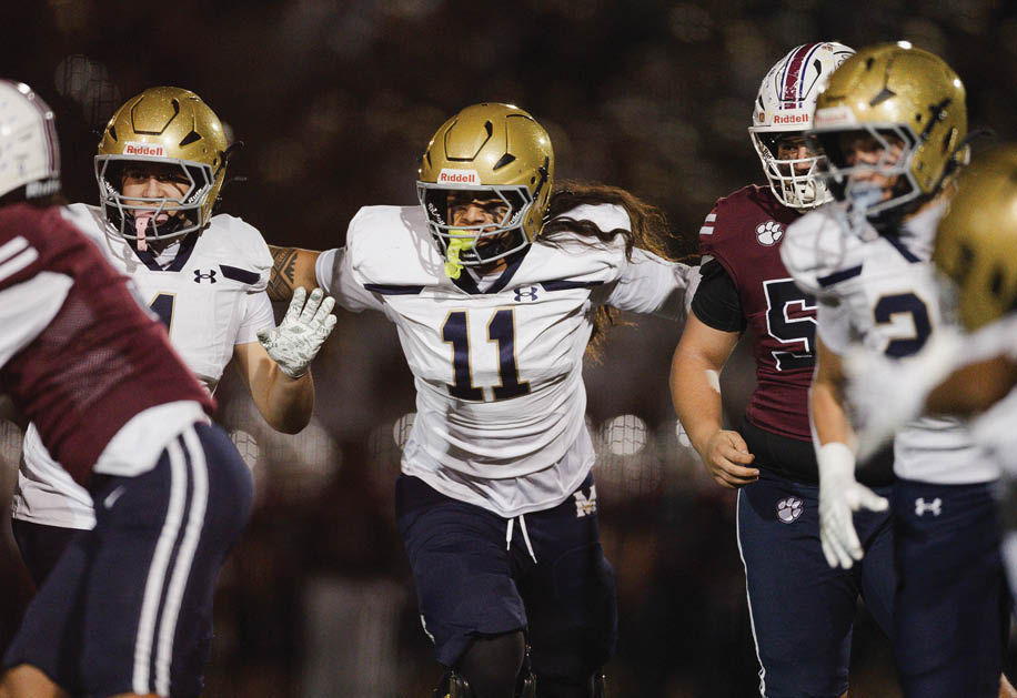 People in football uniforms are actively engaged in a game, with one player in a white jersey and gold helmet running forward while others in maroon and white jerseys prepare to block or tackle. Description generated by AI