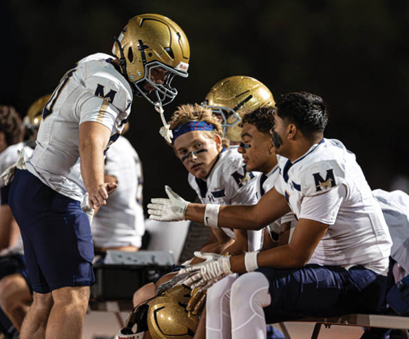 People wearing football uniforms and helmets are sitting on a bench, with one person standing and engaging in conversation with the others during a game. Description generated by AI