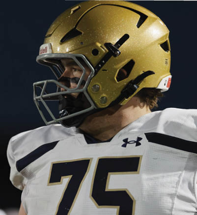 A person wearing a gold football helmet and a white jersey with the number 75 is looking to the side against a dark background. Description generated by AI