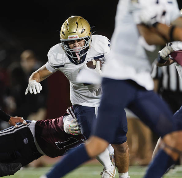Two people in football uniforms are engaged in a play on the field with one person pulling the leg of another who is holding the ball while a third person approaches. Description generated by AI
