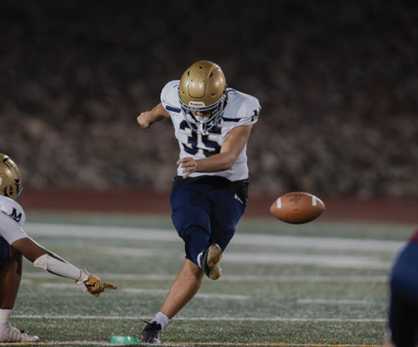 A person wearing a white and blue football uniform with a gold helmet is kicking a football on a field while another person in similar attire points towards the ball. Description generated by AI