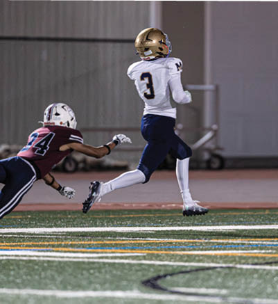 A person in a white and gold football uniform is running on a field while another person in a maroon and white uniform dives attempting to catch them. Description generated by AI