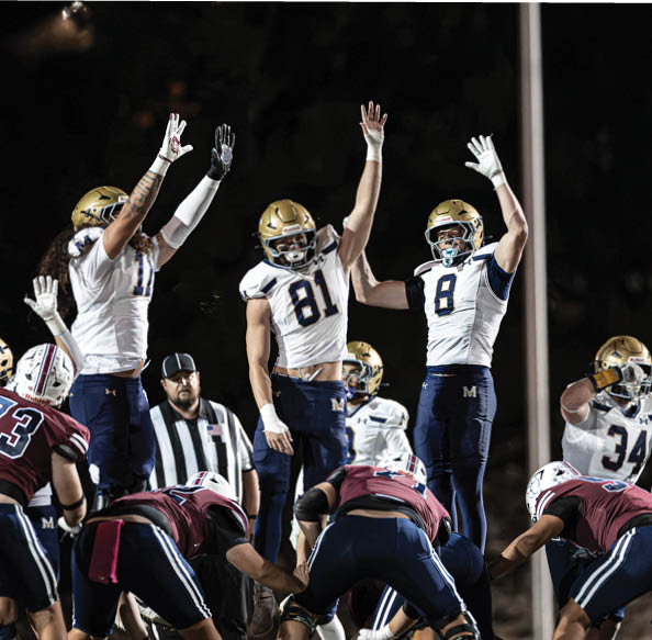People in football uniforms are positioned on a field at night, with one person raising both arms while others are crouched in a ready stance. Description generated by AI