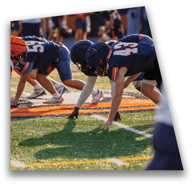 A person in football gear and helmet is crouched on a field with other players lined up in the background during a game. Description generated by AI
